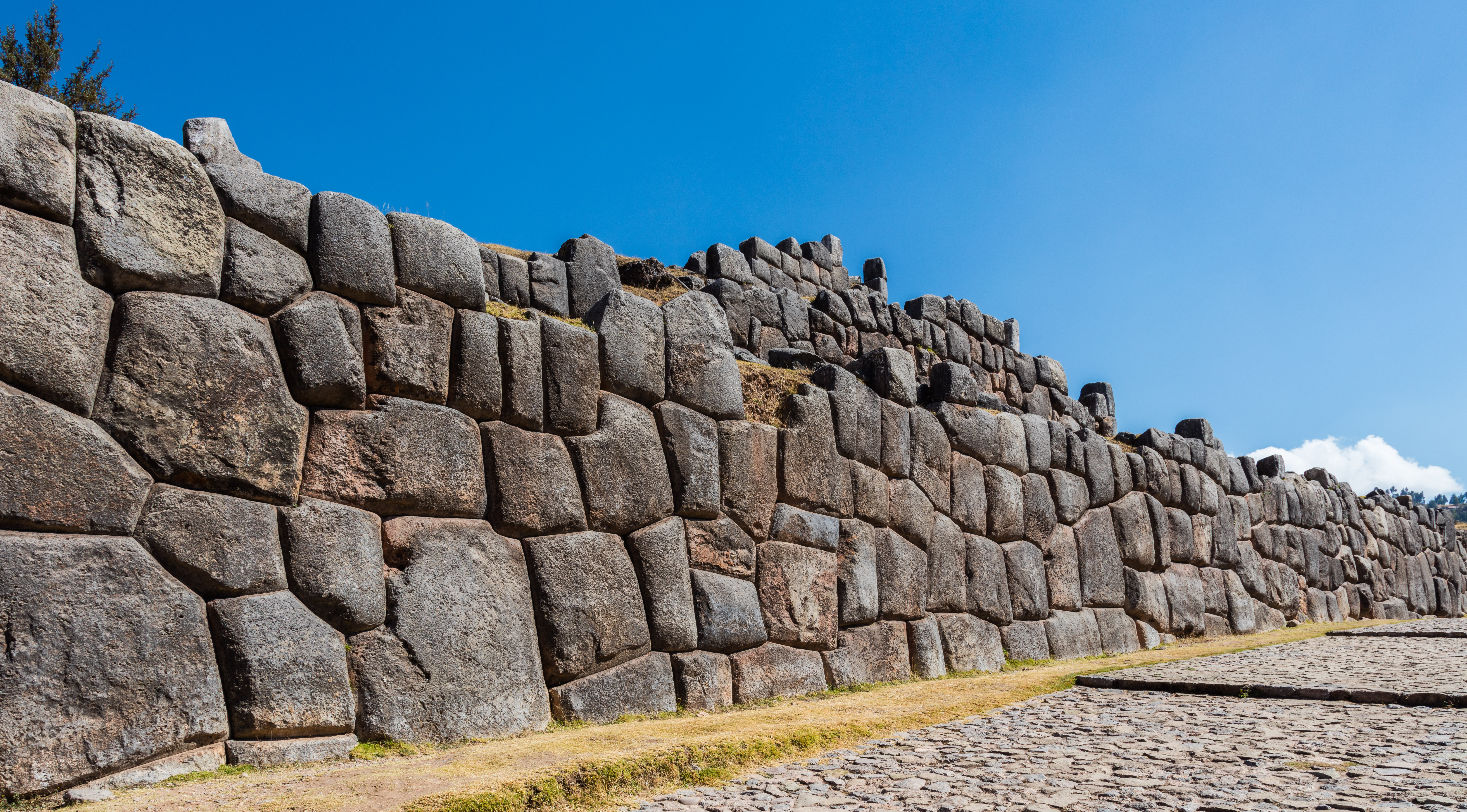 The lower outer wall at Sacsayhuamán above Cusco: massive polygonal andesite blocks fitted without mortar, many of them with seven or more sides, weathered grey-gold under a deep blue Andean sky.