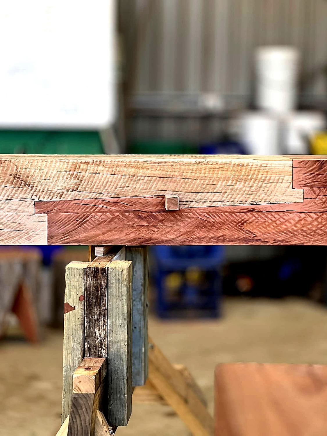 Close-up photograph of a finished Daimochi Tsugi Japanese scarf joint in timber, with a square wooden lock peg visible at the centre of the joint and the stepped complementary cuts running across the beam.