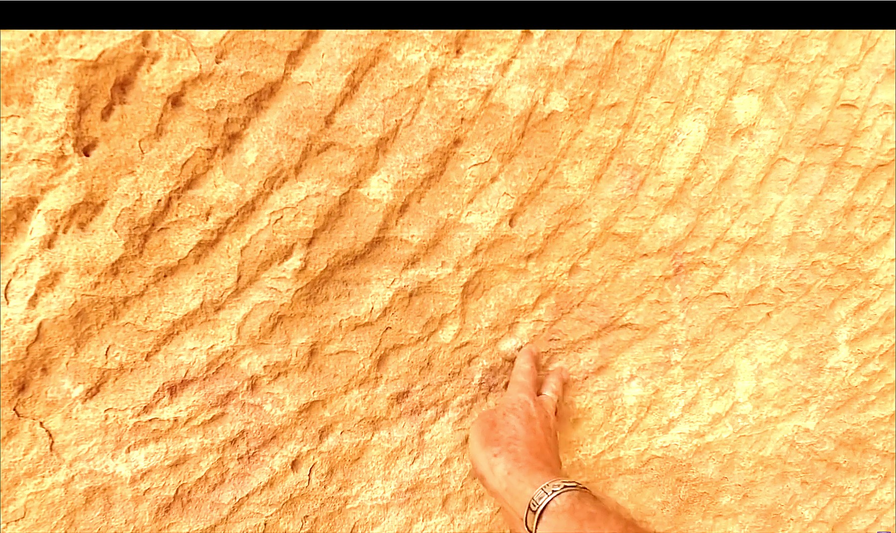 A hand on a sandstone wall at Little Petra showing diagonal raking tool marks crossing the rock face.